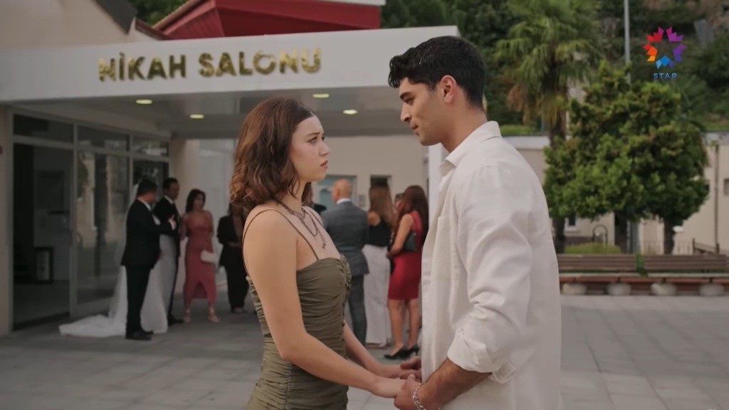 Seyran stoi naprzeciwko Sinana. Oboje trzymają się za ręce. W tle widać salę weselną.