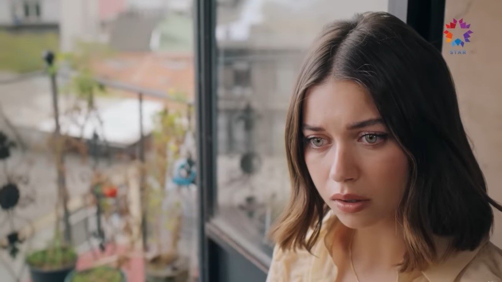 Seyran jest wstrząśnięta. Siedzi przy otwartych drzwiach balkonowych. Na balkonie znajdują się kwiaty. W tle widać dachy innych budynków.