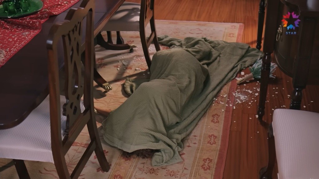 Na podłodze leżą zwłoki, przykryte oliwkowym kocem. Obok znajdują się odłamki porcelany z rozbitego wazonu.