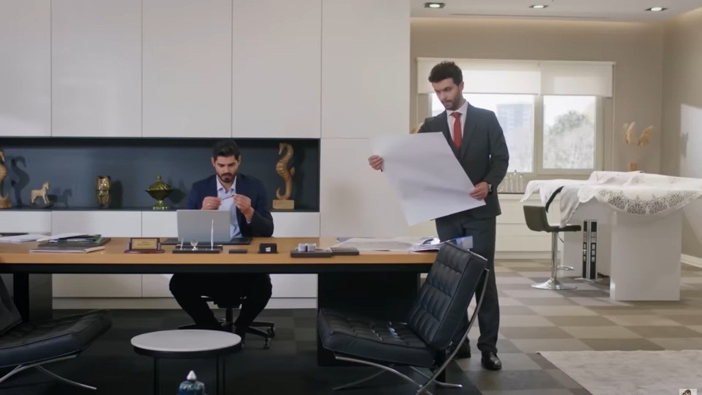 Cihan siedzi za biurkiem. Obok stoi Engin, trzymając w rękach duży arkusz papieru.