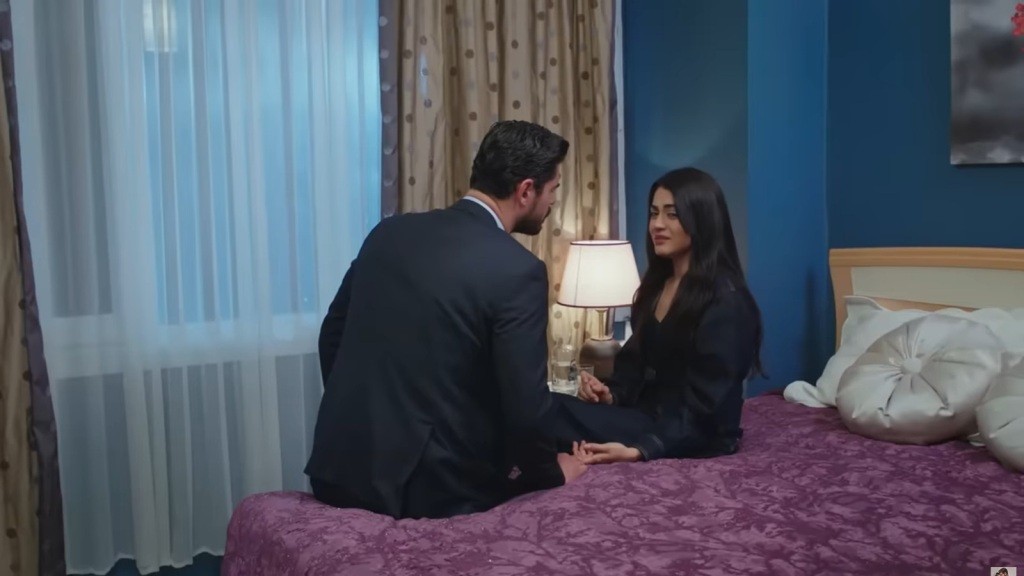 Cihan siedzi na łóżku obok Hancer. Pokój oświetlony jest ciepłym światłem lampy nocnej.