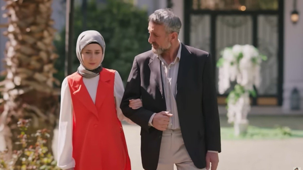 Mężczyzna w eleganckim garniturze prowadzi młodą kobietę ubraną w kolorową sukienkę i chustę na głowie.