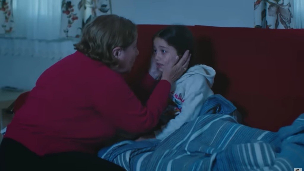 Starsza kobieta kuca przy kanapie i obejmuje policzki przestraszonej Mine.