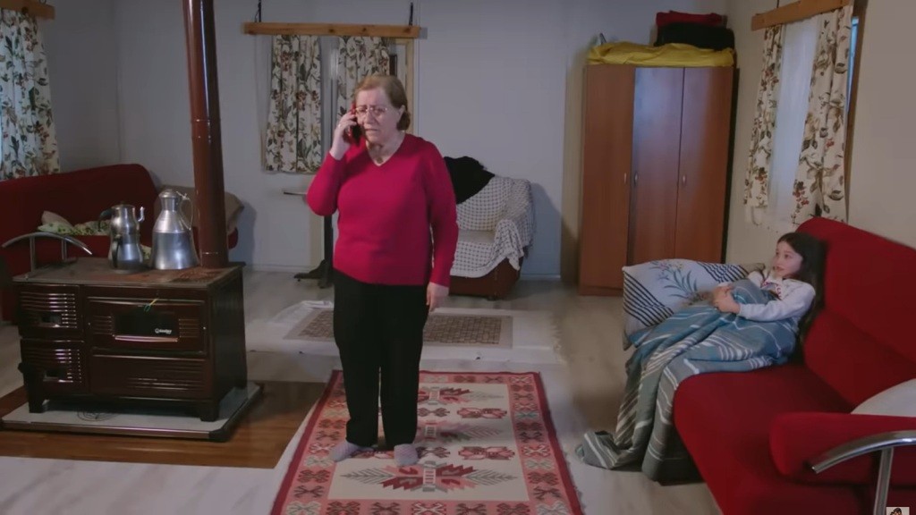 Mine siedzi na kanapie, otulona kocem. Starsza kobieta stoi na środku pokoju i rozmawia przez telefon.