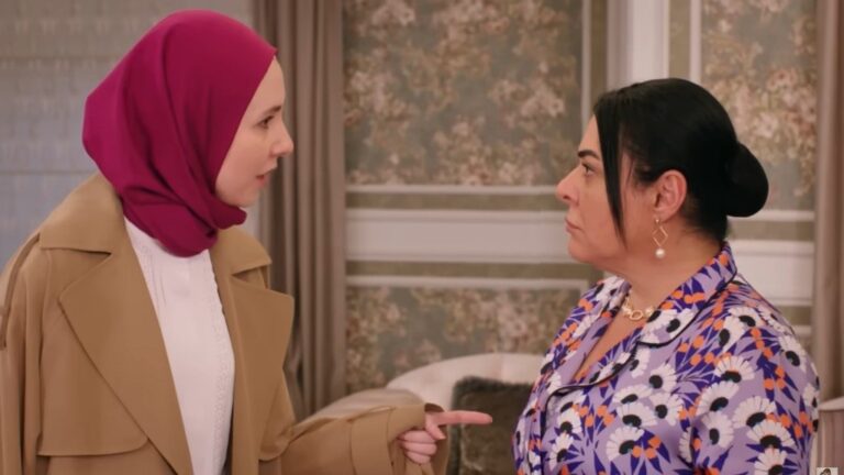 Intensywna rozmowa między dwiema kobietami, gdzie jedna, w kolorowej chustce, wyraża swoje zdanie z determinacją, a druga, w eleganckiej odzieży, słucha z uwagą. Ich napotkane spojrzenia i gesty podkreślają napiętą atmosferę, a sytuacja wydaje się być kluczowa w ich relacji. Scena odzwierciedla emocjonalne zmagania i różnice perspektyw.