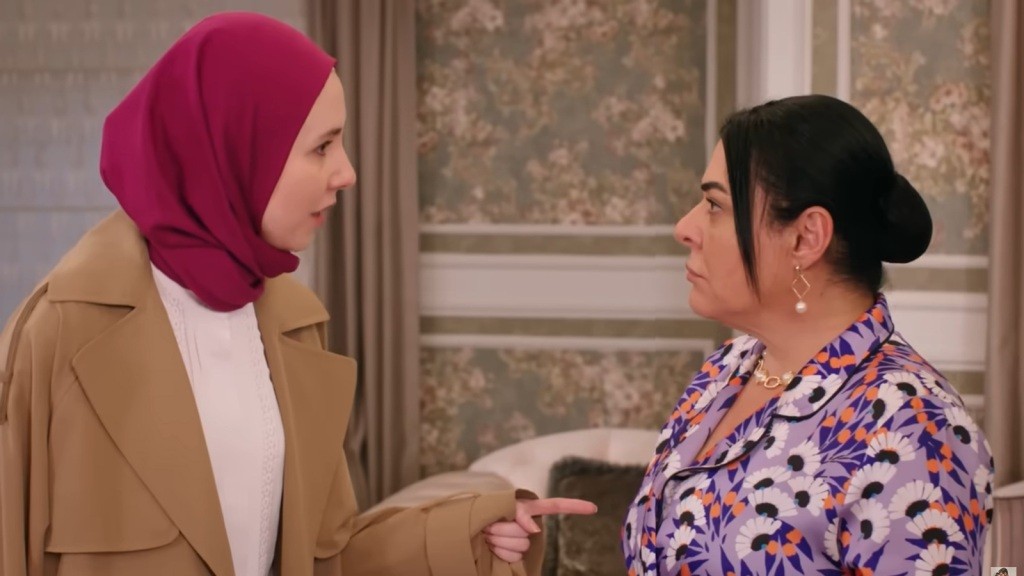 Intensywna rozmowa między dwiema kobietami, gdzie jedna, w kolorowej chustce, wyraża swoje zdanie z determinacją, a druga, w eleganckiej odzieży, słucha z uwagą. Ich napotkane spojrzenia i gesty podkreślają napiętą atmosferę, a sytuacja wydaje się być kluczowa w ich relacji. Scena odzwierciedla emocjonalne zmagania i różnice perspektyw.