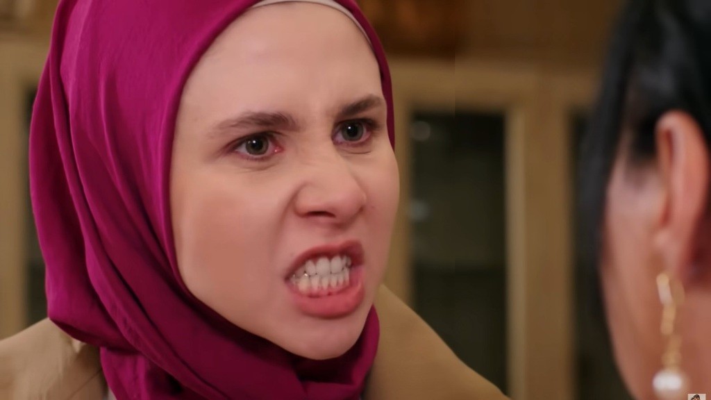 Ekspresyjna scena przedstawiająca kobietę w chustce, wyrażającą silne emocje i złość. Jej intensywne spojrzenie i napięte mięśnie twarzy tworzą dramatyczny moment konfrontacji. W tle widać drugą kobietę, która wydaje się słuchać z niepewnością. Ta emocjonalna chwila ukazuje konflikt oraz napięcie między postaciami.