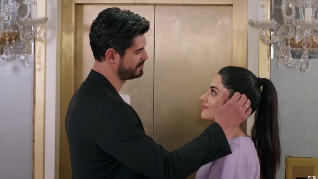 Urocza scena przedstawiająca mężczyznę i kobietę, którzy wymieniają czułe spojrzenia, stojąc przed drzwiami windy. Mężczyzna delikatnie dotyka włosów kobiety, co podkreśla intymność momentu. Ich wyrazy twarzy mówią o bliskości i wzajemnym zrozumieniu, tworząc atmosferę romantycznego napięcia.