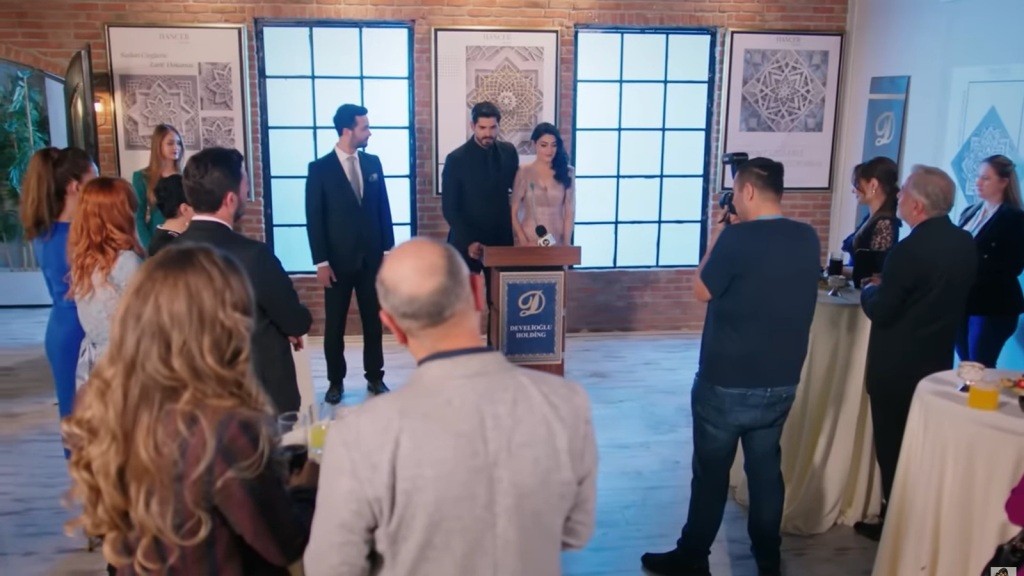 Wydarzenie promujące nową kolekcję dywanów. Cihan i Hancer stoją przy podium. Wokół zgromadzeni są goście.