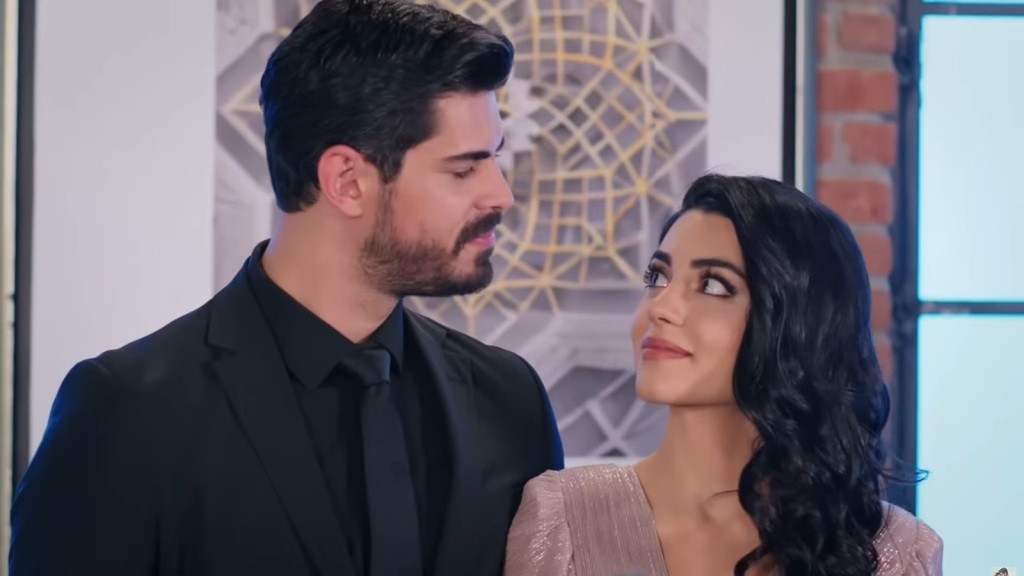 Na zdjęciu Cihan i Hancer dzielą moment intymności, patrząc sobie głęboko w oczy. Oboje ubrani w eleganckie stroje, ich wyraz twarzy wyraża uczucia miłości i zaufania. Tło z nowoczesnymi wzorami dodaje scenie stylowego charakteru.