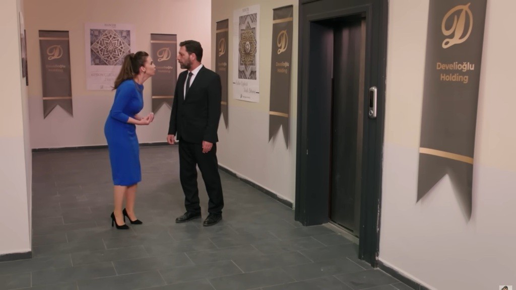 Scena w biurowym korytarzu, gdzie postacie Beyza i Nusret prowadzą emocjonalną rozmowę, odzwierciedlając napięcie i dramat ich relacji. W tle widoczne są eleganckie elementy wystroju z logiem firmy Develioglu Holding.