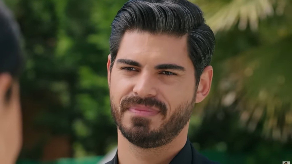 Zbliżenie na mężczyznę o wyrazistej urodzie, który z uśmiechem i pełnym ciepła spojrzeniem wyraża zainteresowanie i otwartość. Jego stylowy wygląd i pewność siebie przyciągają uwagę, tworząc atmosferę przyjacielskiej rozmowy.