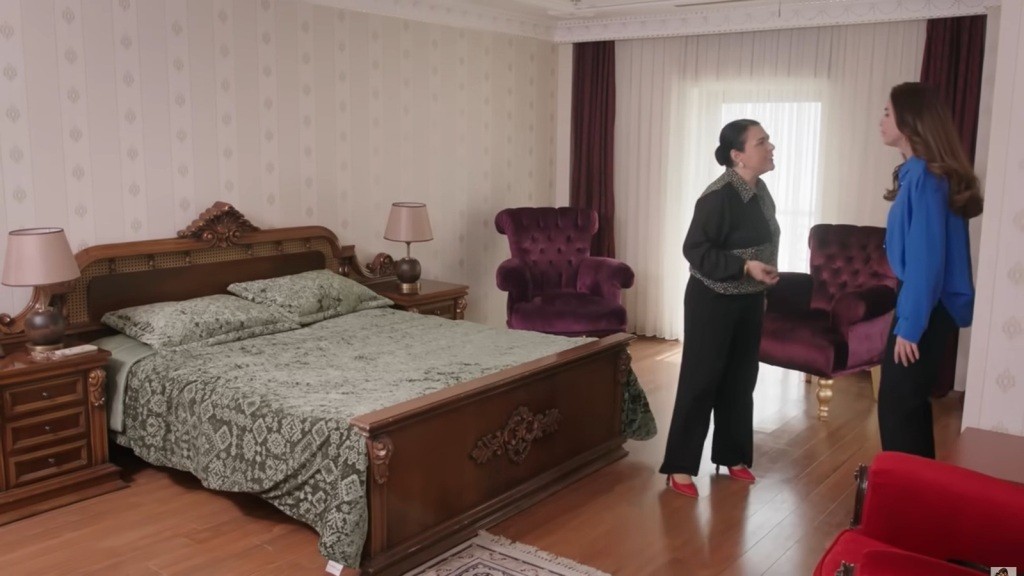 Scena z eleganckiego wnętrza, w którym dwie kobiety prowadzą intensywną rozmowę. W tle widoczna jest przestronna sypialnia z ozdobnym łóżkiem i stylowymi fotelami, co nadaje atmosferę luksusu i napięcia.