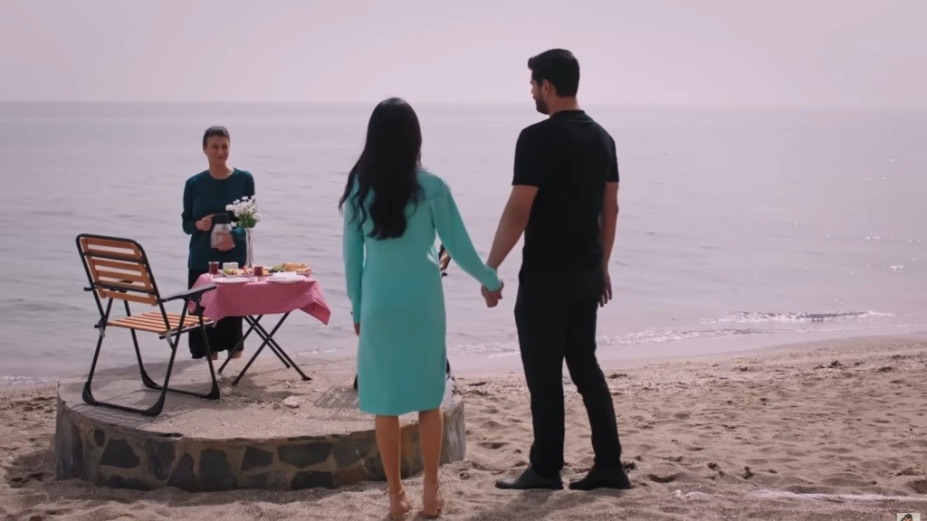 Para trzymająca się za ręce nad brzegiem morza, stolik z posiłkiem na plaży i osoba witająca ich na tle spokojnego oceanu.