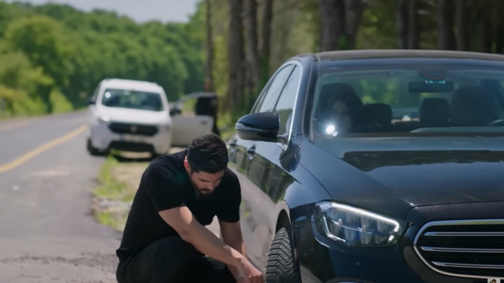 Cihan kuca przy prawym przednim kole czarnej limuzyny. Z zaparkowanego z tyłu białego samochodu wysiada kierowca.
