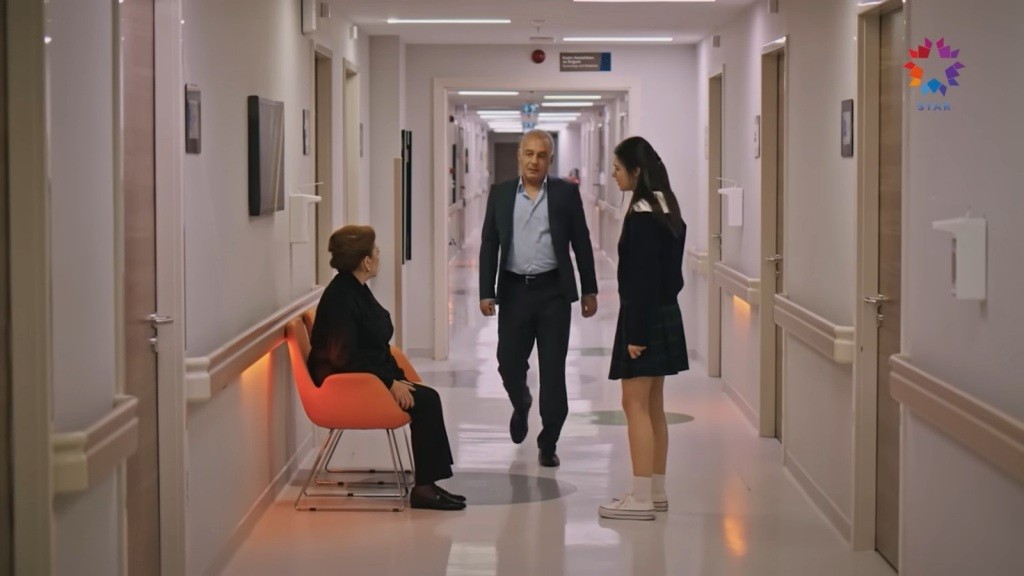 Ayla siedzi na pomarańczowym krzesełku. Na przeciwko niej stoi Hazal. Mumtaz idzie w ich stronę korytarzem.
