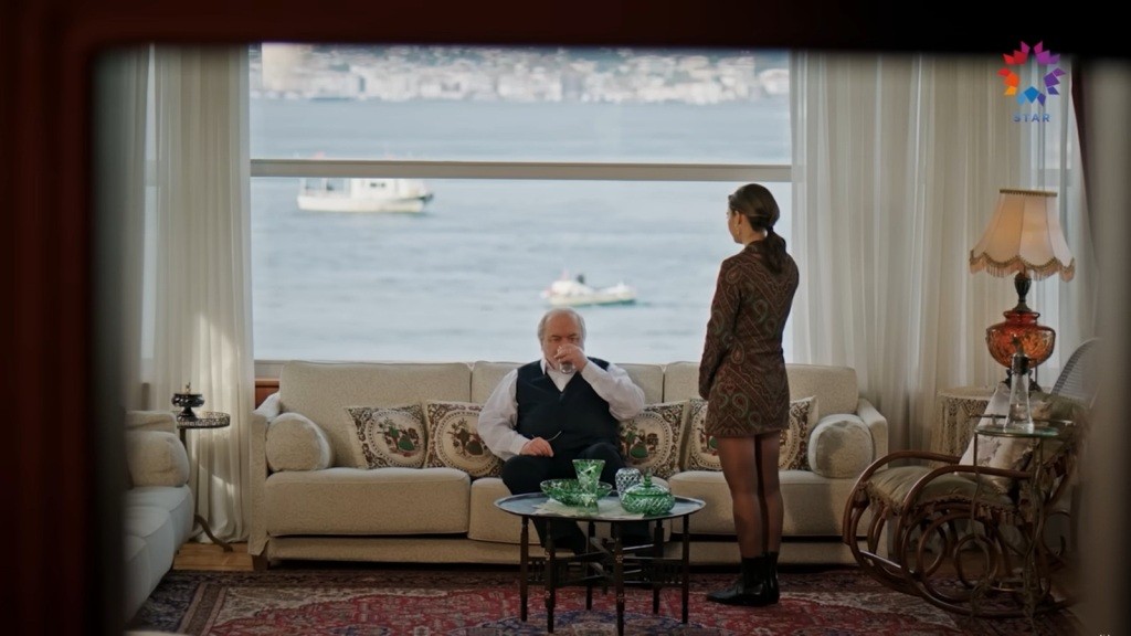 Seyran stoi przed Halisem, który siedzi na kanapie i pije wodę.