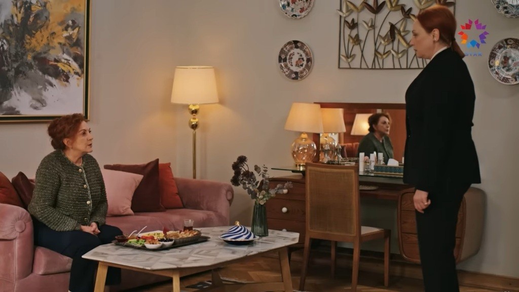 Ayla siedzi na kanapie. Ifakat stoi naprzeciwko niej.