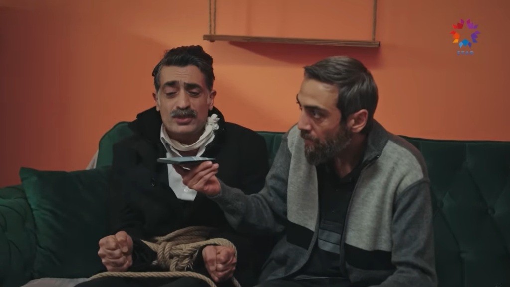 Związany Kazim siedzi na kanapie. Sevki siedzi obok niego i trzyma telefon przy jego ustach.