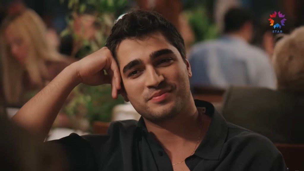 Ferit uśmiecha się lekko, opierając głowę na dłoni.