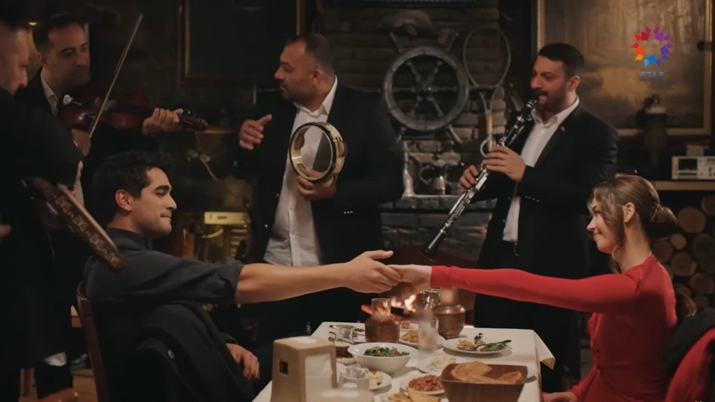 Ferit i Seyran chwytają się za ręce. Obok ich stolika grają muzykanci.