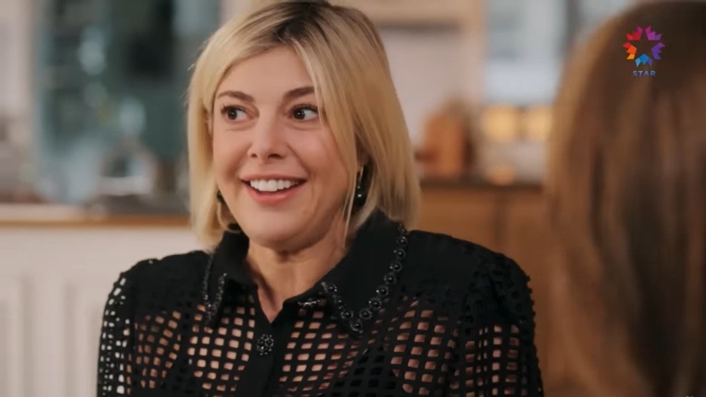 Gulgun ma na sobie czarną, ażurową bluzkę. Jej wyraz twarzy jest pełen radości i zaangażowania.