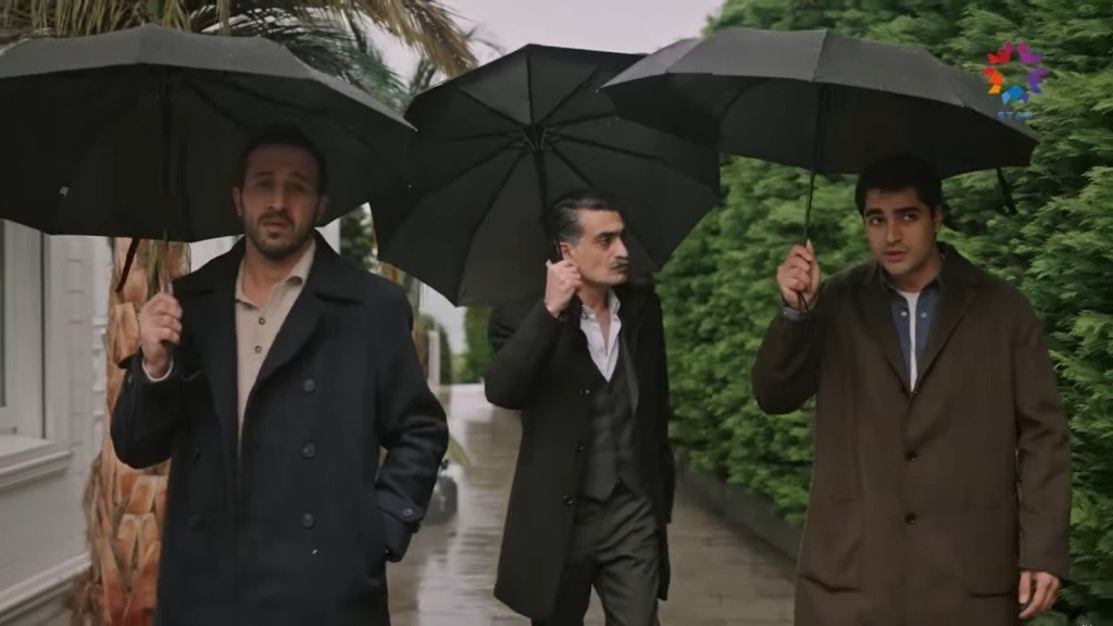 Abidin, Kazim i Ferit idą chodnikiem w deszczu. Każdy trzyma czarny parasol. Ubrani są w płaszcze i otoczeni zielenią.