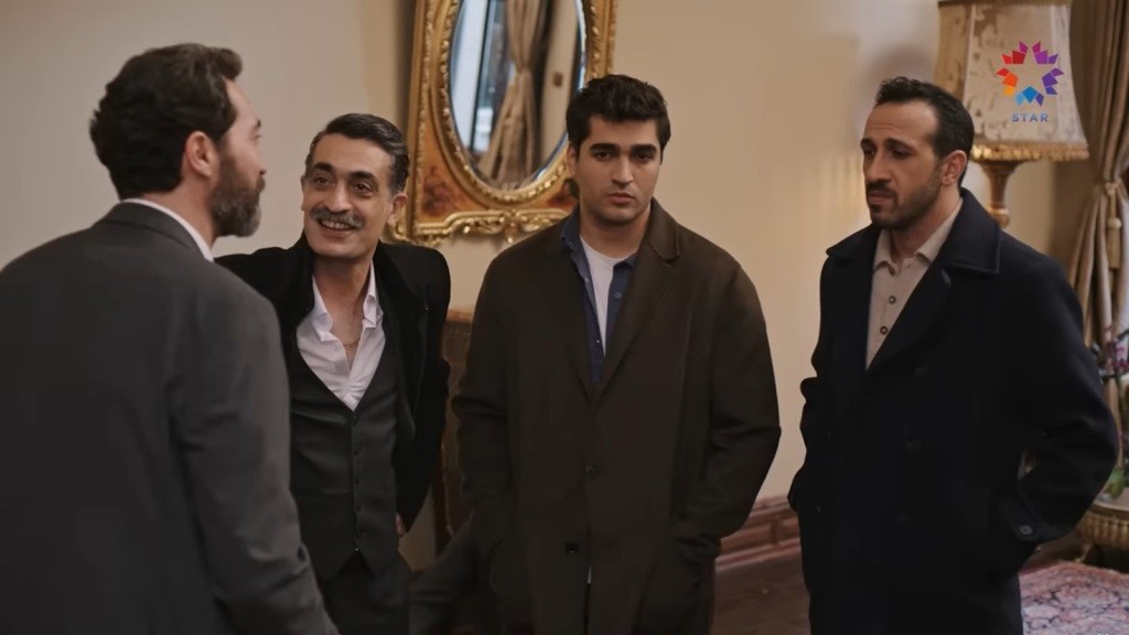 Czterech mężczyzn rozmawia w eleganckim wnętrzu; jeden zwrócony tyłem, trzej pozostali stoją obok siebie przy ozdobnym lustrze.