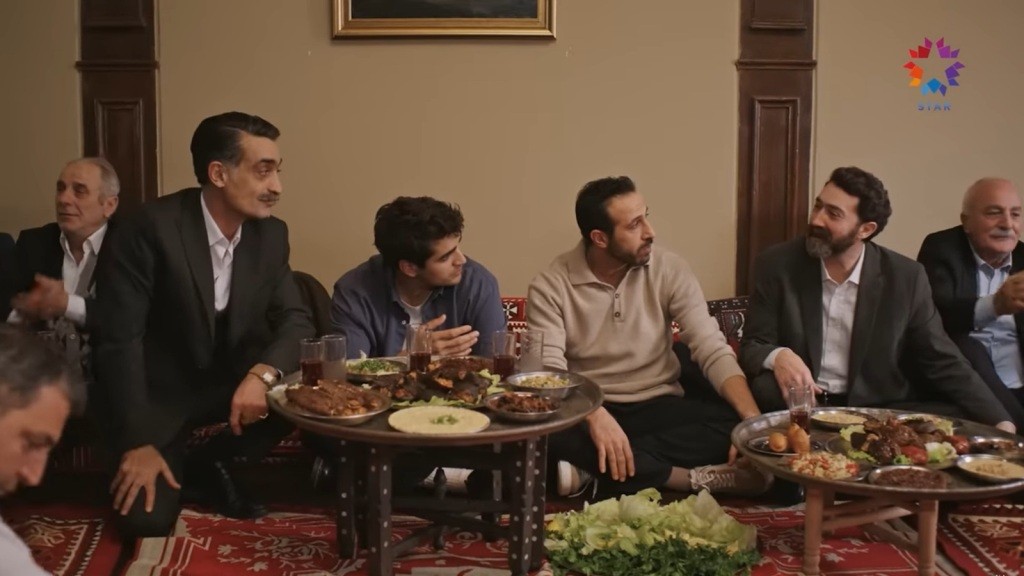 Mężczyźni siedzą razem przy niskich stolikach z potrawami podczas sıra gecesi. Rozmowa i wspólne posiłki w przytulnym, tradycyjnym wnętrzu.