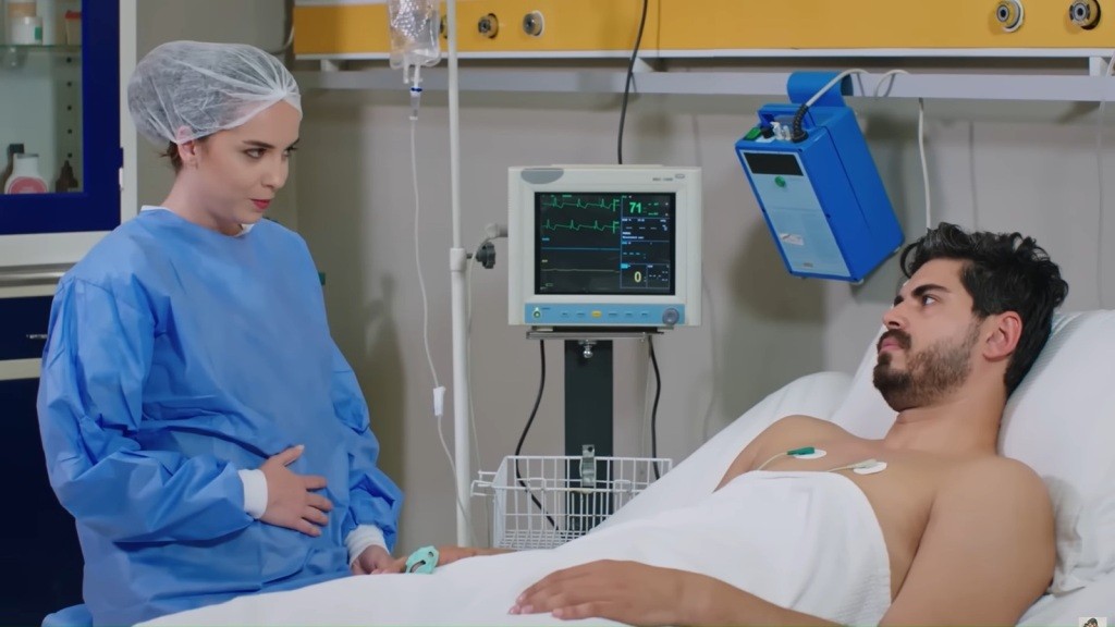 Beyza siedzi przy łóżku Cihana w ubraniu ochronnym. Prawą dłoń przykłada do swojego brzucha i z lekkim uśmiechem patrzy na Cihana.