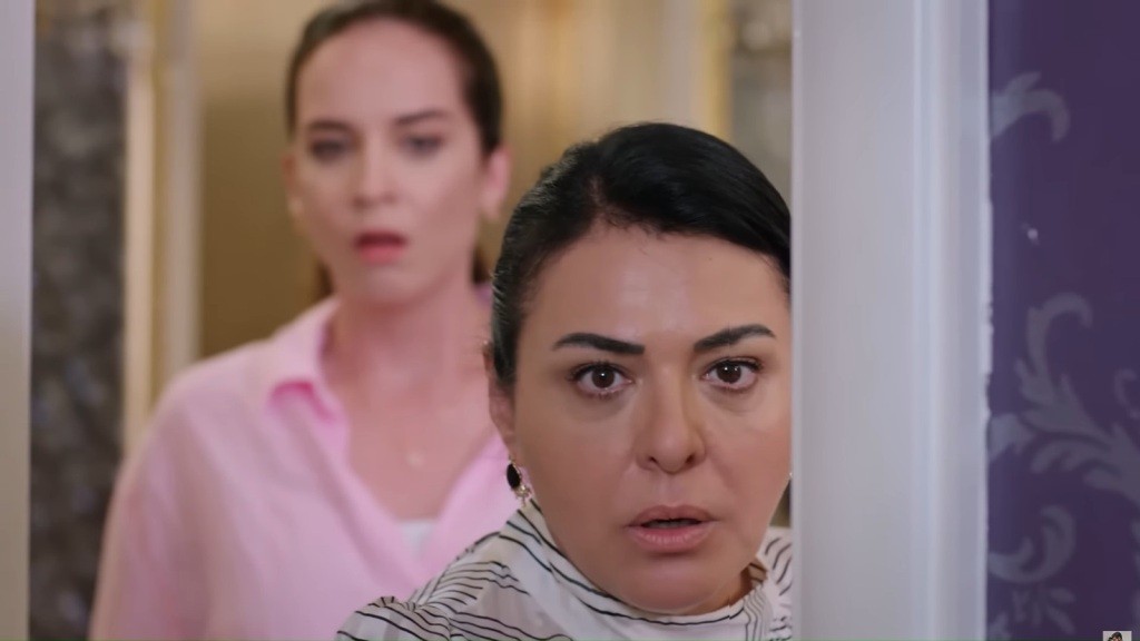 Beyza i Mukadder stoją przed uchylonymi drzwiami, zaglądając do środka pokoju. Obie mają zszokowane wyrazy twarzy.