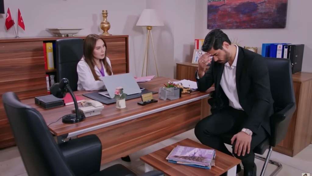 Cihan siedzi z pochyloną głową przy biurku Yasemin.