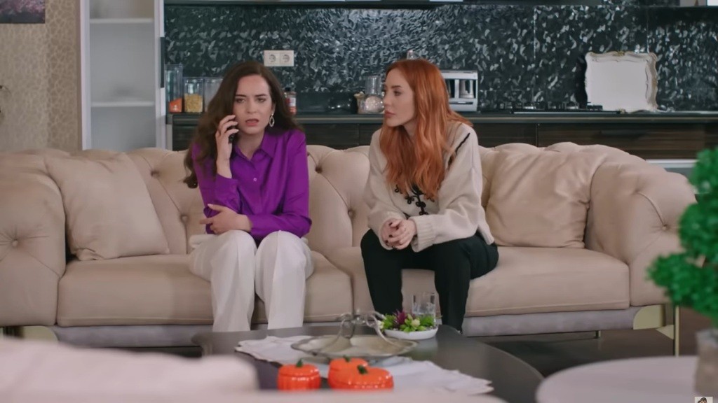 Beyza i Yonca siedzą obok siebie na kanapie. Beyza rozmawia przez telefon, a Yonca przysłuchuje się uważnie.