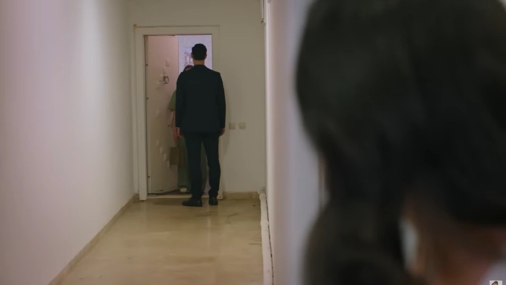 Cihan stoi przed drzwiami mieszkania, które otworzyła mu jakaś kobieta. Na pierwszy planie widać zarys innej postaci, która ich obserwuje.