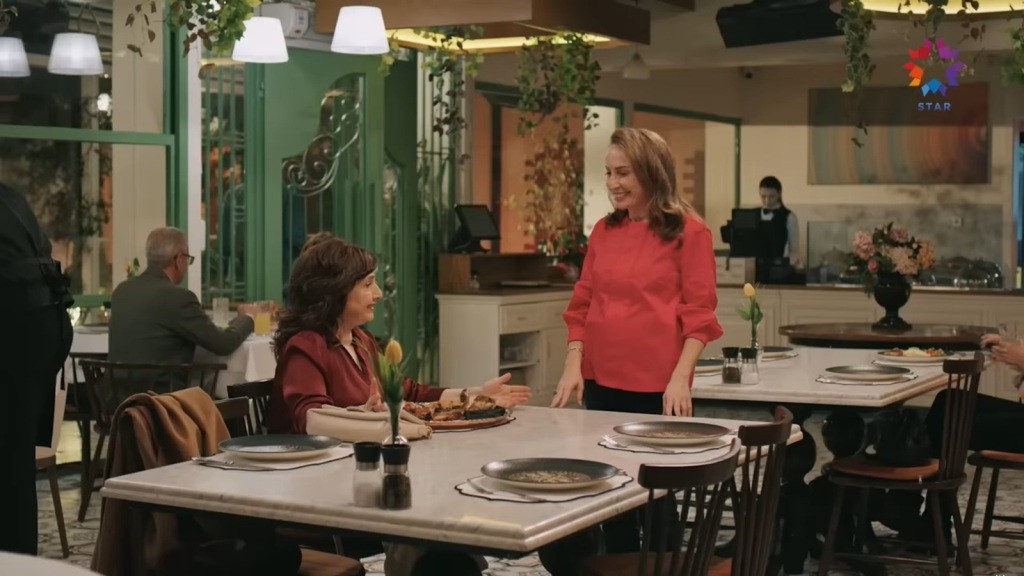 Scena w restauracji. Cicek siedzi przy stole, a Esme stoi tuż obok i uśmiecha się do niej.