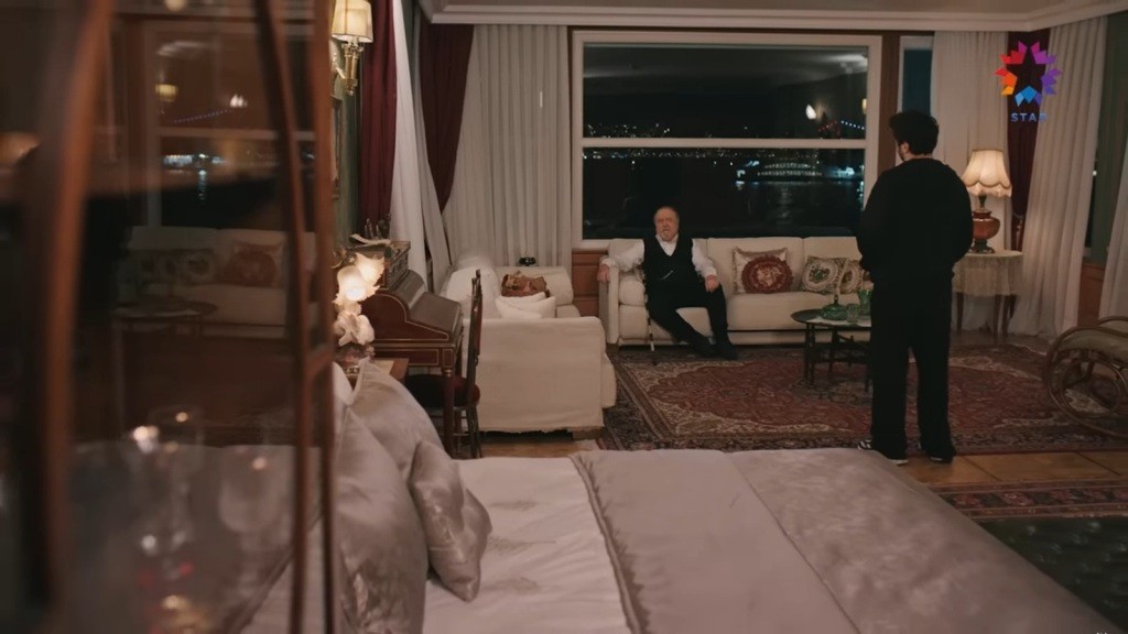 Hali siedzi wygodnie na kanapie. Ferit stoi przed nim.