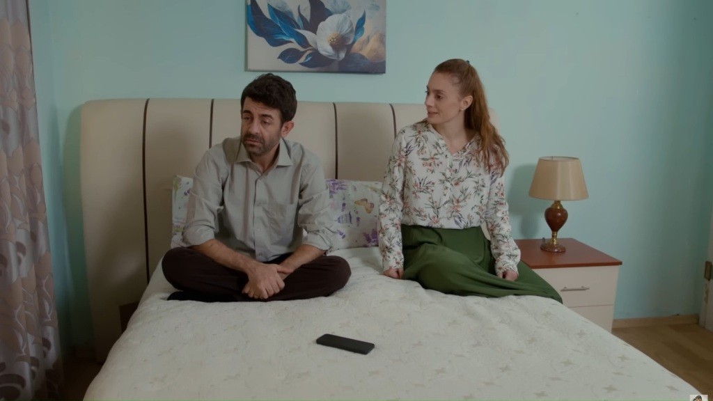 Cemil i Derya siedzą na łóżku. On ze skrzyżowanymi nogami i zgarbiony, ona z troską patrząca na niego. Na łóżku, przed Cemilem, leży także telefon.