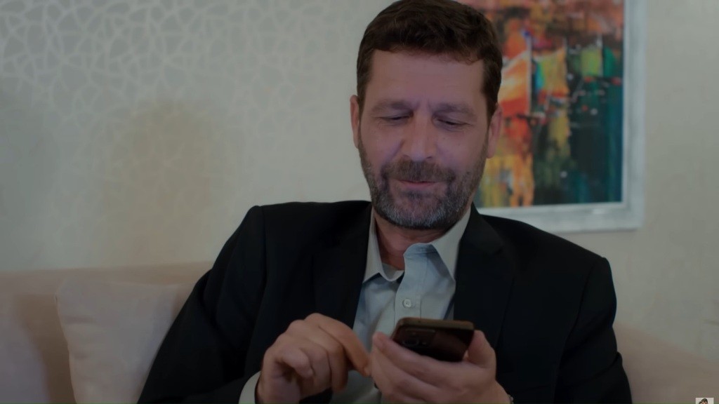 Nusret siedzi na kanapie. Uśmiecha się, patrząc w ekran telefonu.