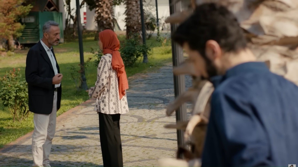 Mahmut i Sinem rozmawiają, stojąc na alejce. Melih ukrywa się za pniem palmy.
