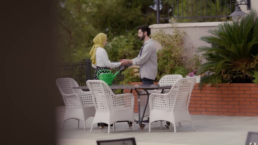 Melih i Sinem stoją w oddali ze splecionymi dłońmi. Przed nimi jest okrągły stół, na nim zielona konewka, a wokół niego białe krzesła.