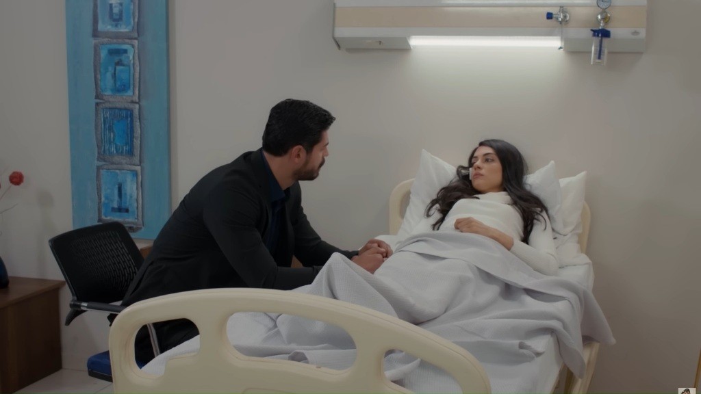 Cihan siedzi przy łóżku Hancer i trzyma ją za rękę.