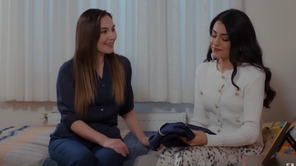 Yasemin i Hancer siedzą obok siebie na łóżku. Yasemin uśmiecha się, a Hancer trzyma w rękach granatową, złożoną w kostkę piżamę.