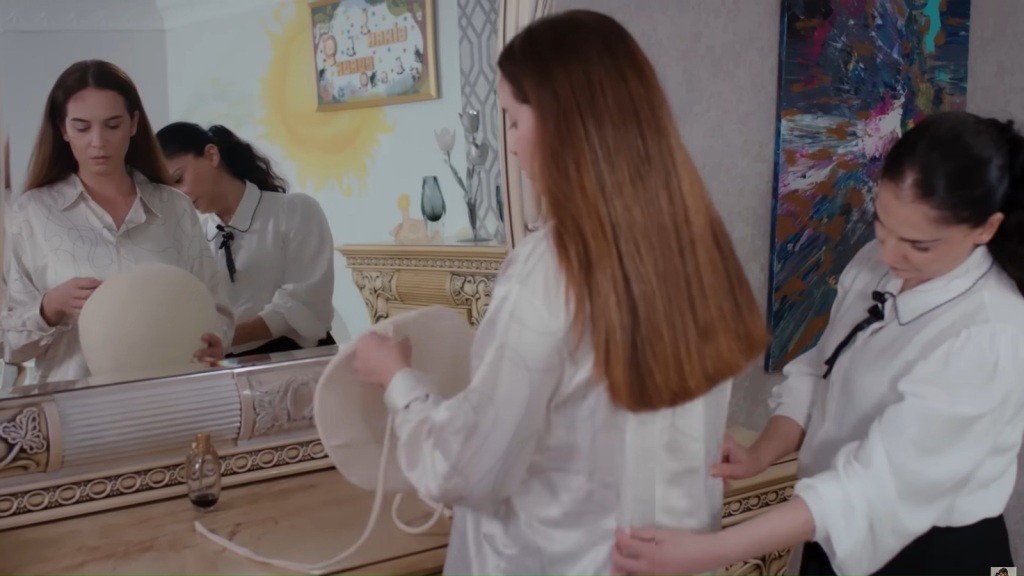 Beyza stoi przed lustrem, trzymając w rękach sztuczny brzuch ciążowy. Gulsum poprawia jej koszulę.