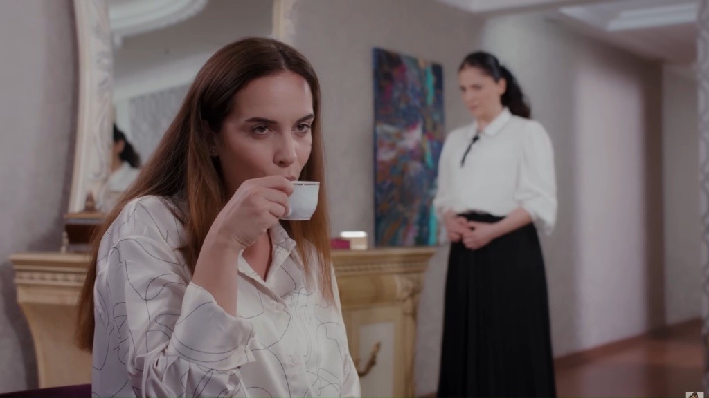 Beyza siedzi i pije kawę. W tle widać stojącą Gulsum.