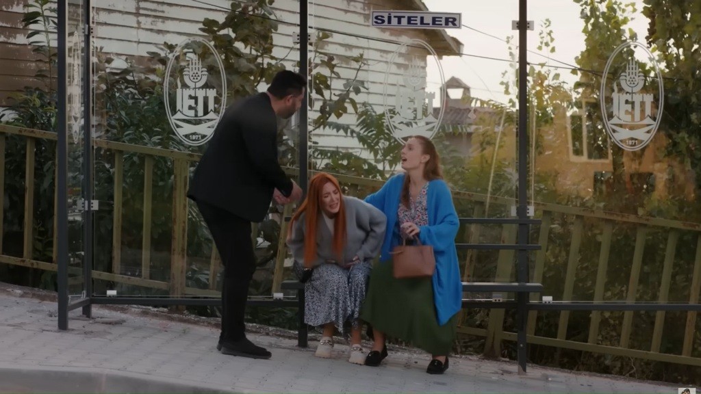 Yonca zwija się z bólu na przystanku. Obok niej siedzi Derya, a nad nimi stoi mężczyzna.