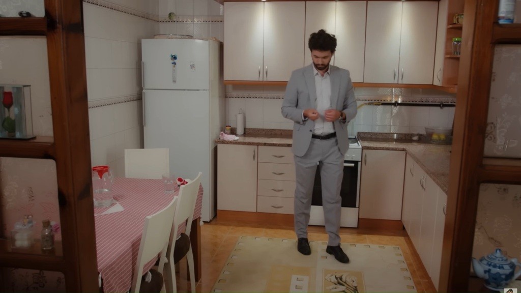 Engin, ubrany w szary garnitur, stoi w kuchni i patrzy w dół; w tle lodówka i stół z kraciastym obrusem.