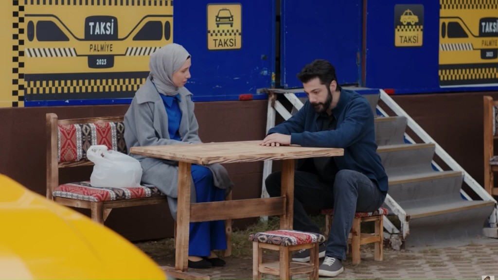 Sinem i Melih siedzą przy drewnianym stoliku przed żółto-niebieską przyczepą.