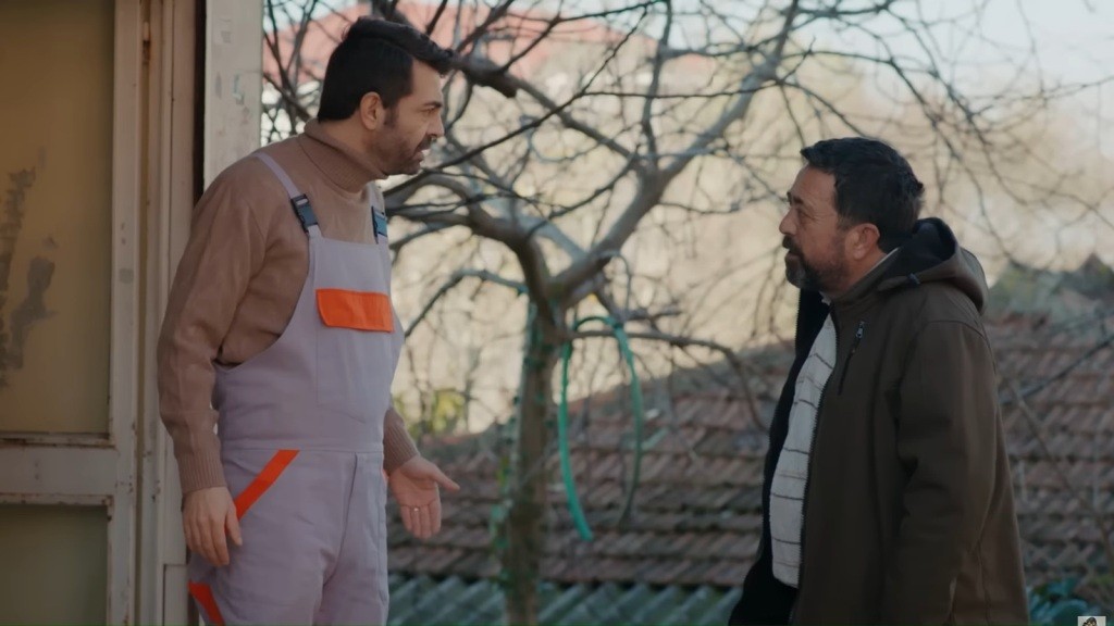 Dwaj mężczyźni prowadzą intensywną rozmowę na zewnątrz domu; jeden w szarym kombinezonie z pomarańczowymi wstawkami.