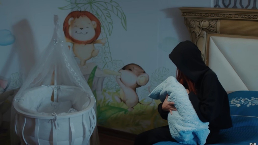 Kobieta w ciemnym kapturze siedzi na łóżku w pokoju dziecięcym, trzymając na rękach owinięte w kocyk niemowlę.