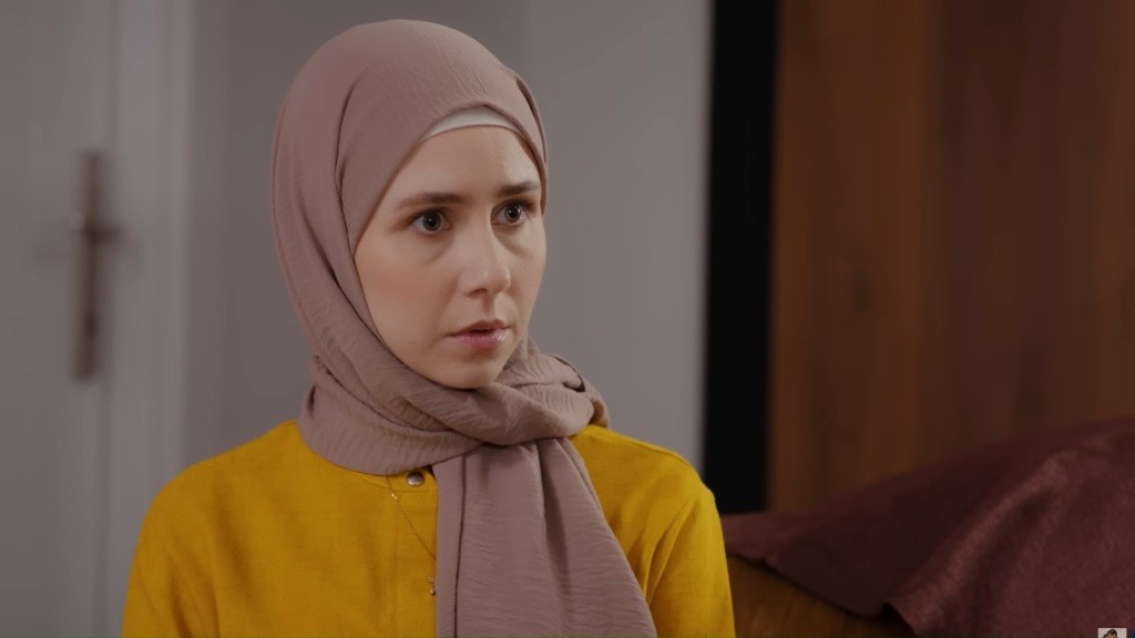 Sinem ma beżową chustę na głowie i zaniepokojony wyraz twarzy.